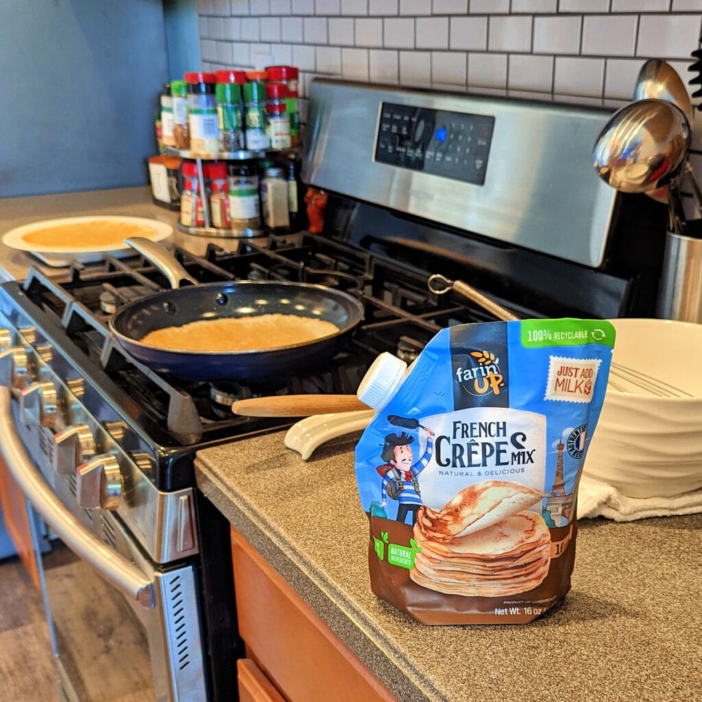 Package of Farin'UP Crepe Mix on countertop, next to stove with crepe cooking in skillet.