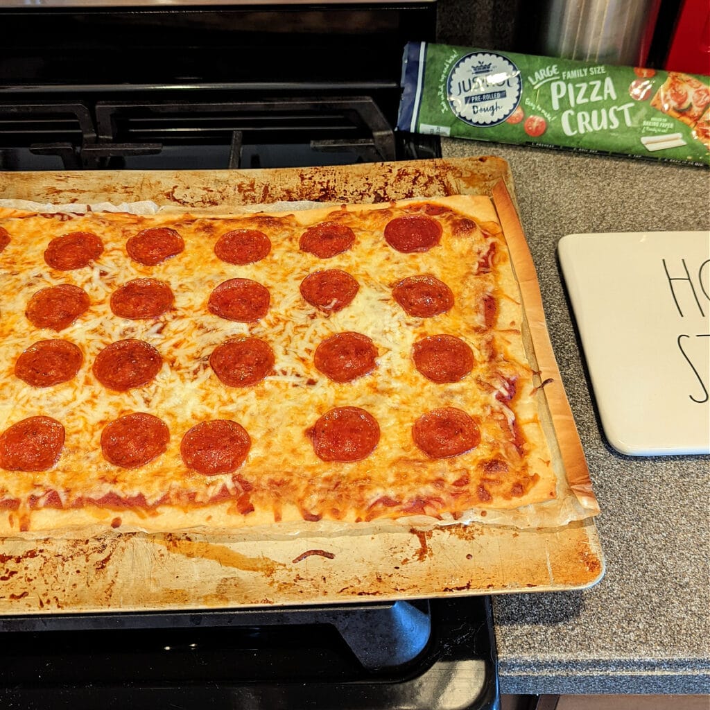 Homemade pepperoni pizza sitting on top of stove, next to Roll-Jus pizza dough packaging.